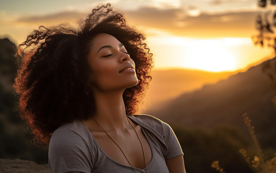 Serene looking woman with eyes closed and head tilted back in front of a sunset representing stress management in recovery